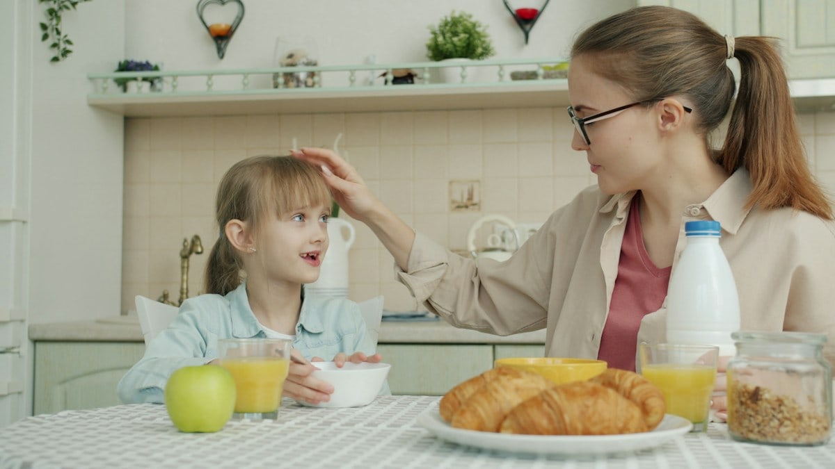 Little girl is having breakfast and talking to mother sitting at table in kitchen in apartment, young woman is touching daughter's hair expressing love and care.